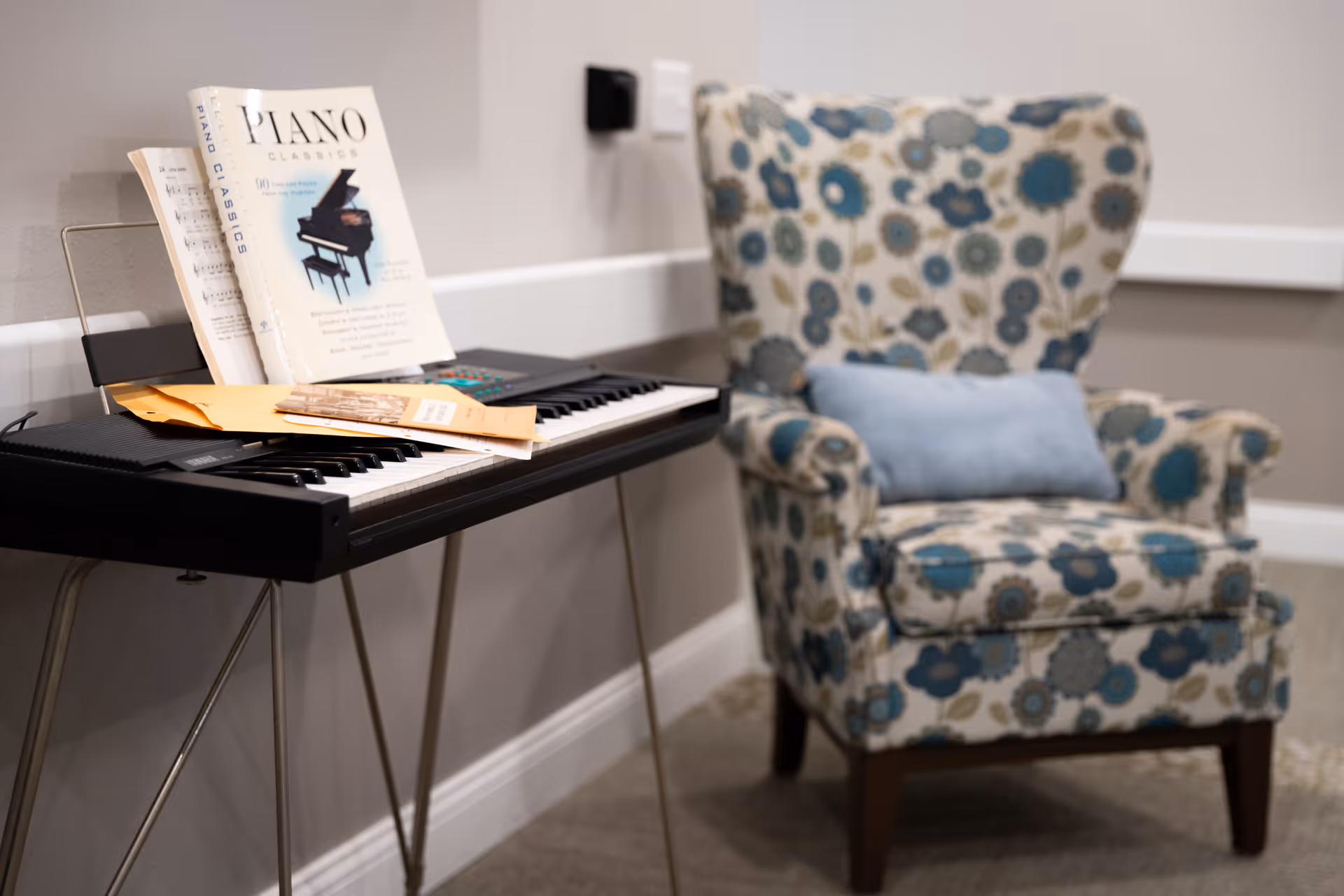 Electronic keyboard with sheet music and envelopes beside a floral patterned armchair with a blue cushion in a cozy interior corner.