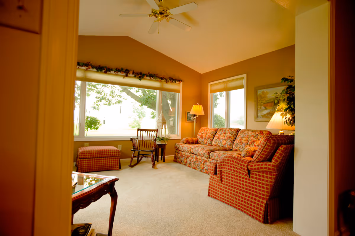A cozy living room with a floral patterned sofa, a matching armchair, a wooden rocking chair, and a cushioned ottoman. The room has large windows letting in natural light, beige carpet, a ceiling fan, and two lamps providing warm lighting. There is a framed picture on the wall and some greenery visible outside the windows.