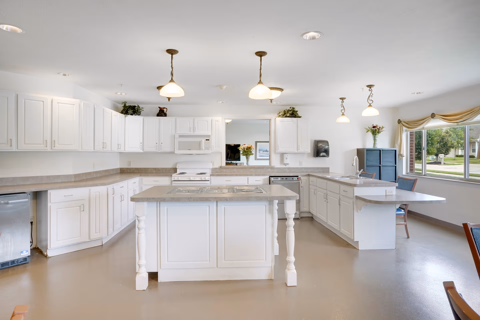 A spacious, bright kitchen with white cabinetry and countertops. The kitchen features a central island with a built-in stovetop, a microwave above the stove, a dishwasher, and a large window letting in natural light. There are pendant lights hanging from the ceiling, and a few decorative plants and flowers placed on the countertops and cabinets.
