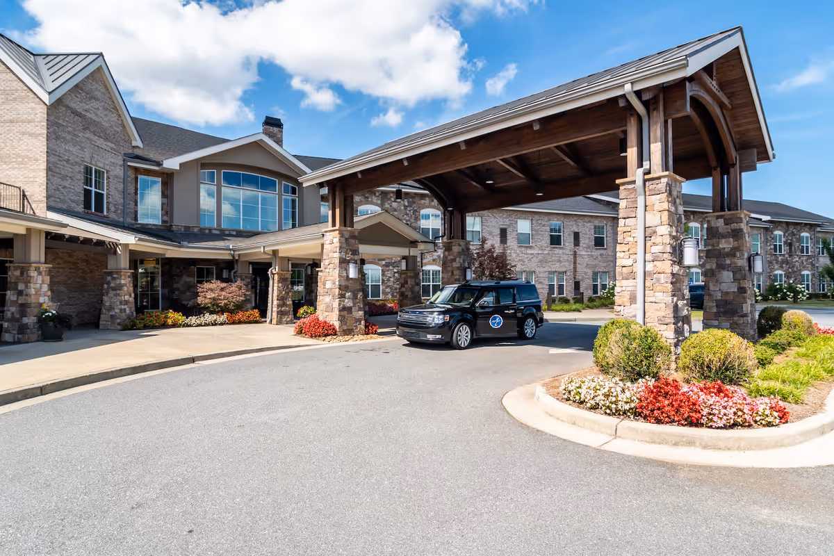Covered porte-cochere entrance of a senior living building with stone pillars, landscaping, and a parked black SUV.