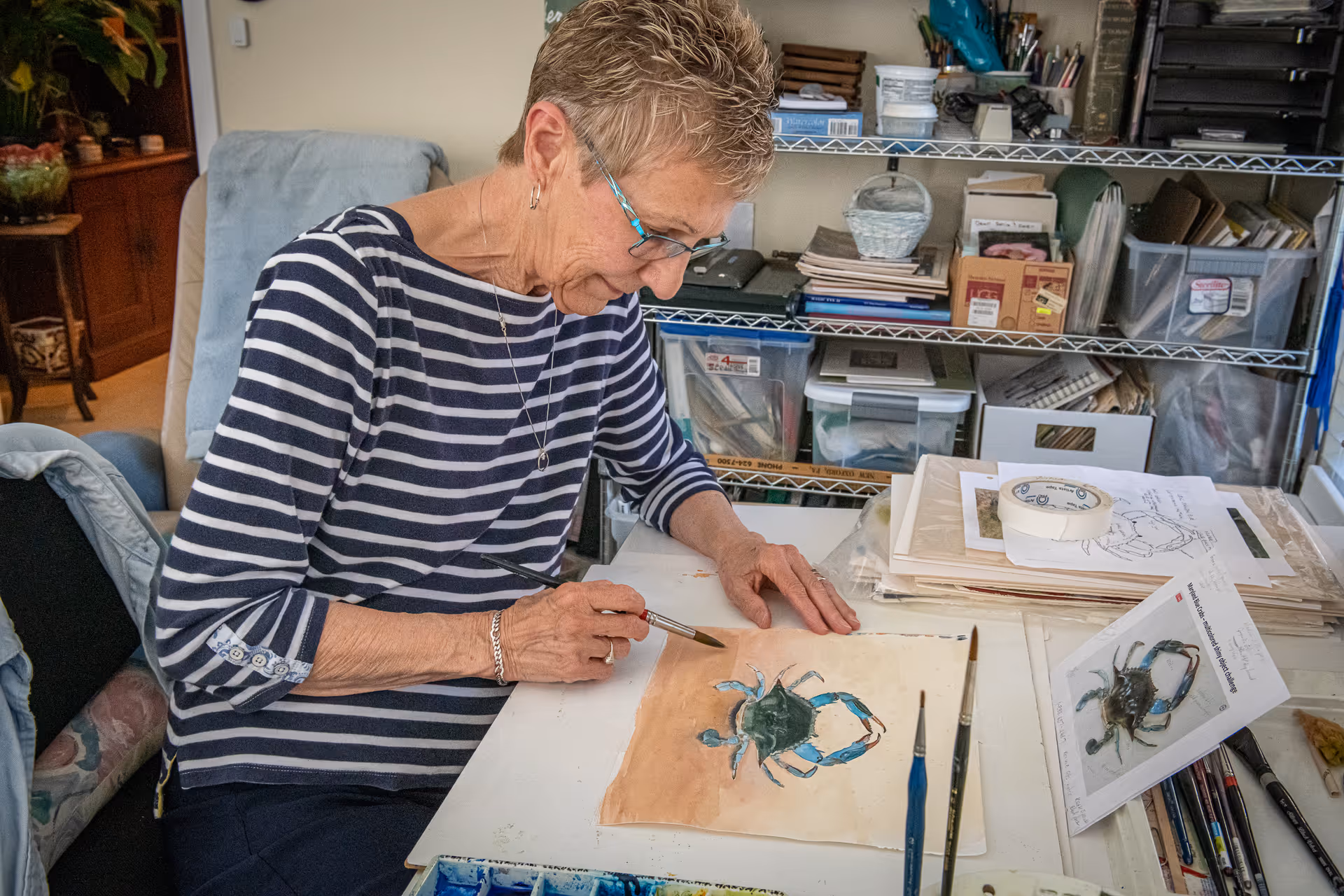 An elderly woman wearing glasses and a striped shirt is sitting at a table painting a picture of a blue crab. The table is cluttered with art supplies, brushes, papers, and a reference photo of a crab. Behind her, there are shelves filled with various art materials and storage boxes.