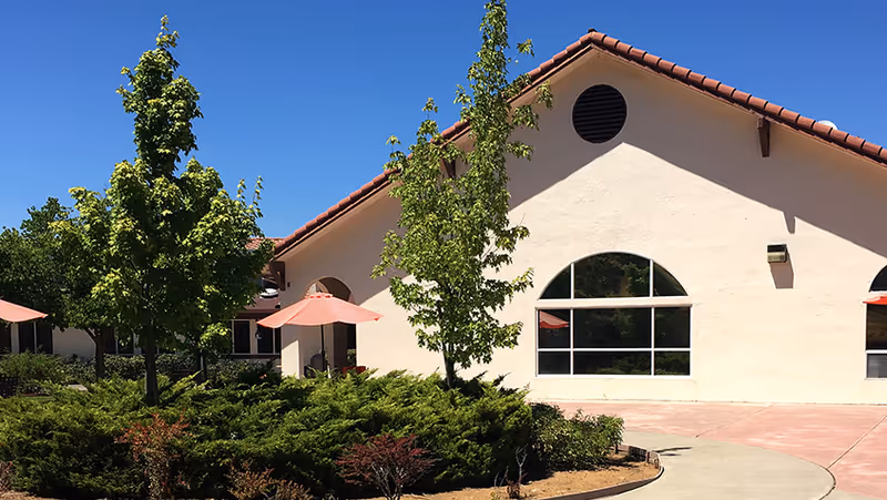 Exterior view of a single-story building with a red tile roof and large arched windows. The foreground features a landscaped area with green shrubs, small trees, and pink patio umbrellas. The sky is clear and blue.
