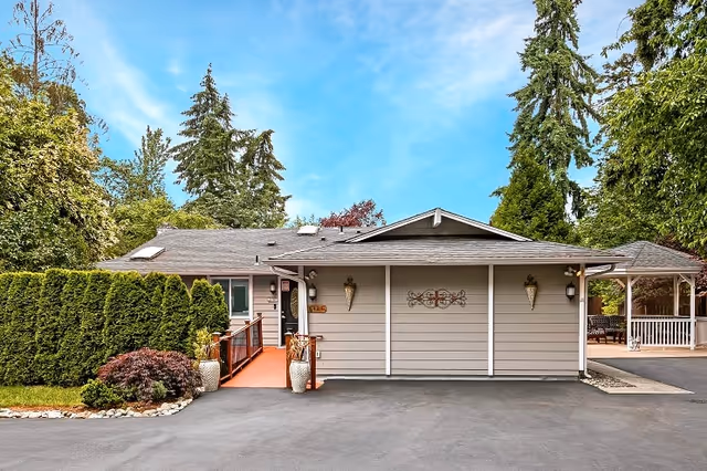 Front exterior view of a single-story senior care home building with a gray facade, a small ramp leading to the entrance, decorative wall sconces, and a covered porch area to the right. The building is surrounded by tall green trees and shrubs under a clear blue sky.