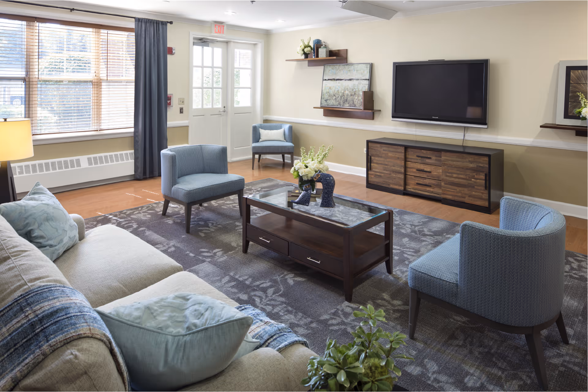 A cozy living room with a beige sofa adorned with blue pillows and a blue throw blanket, three blue upholstered chairs, a wooden coffee table with a glass top and decorative items, a flat-screen TV mounted on the wall above a wooden media console, large windows with blinds and curtains, and a door leading outside.