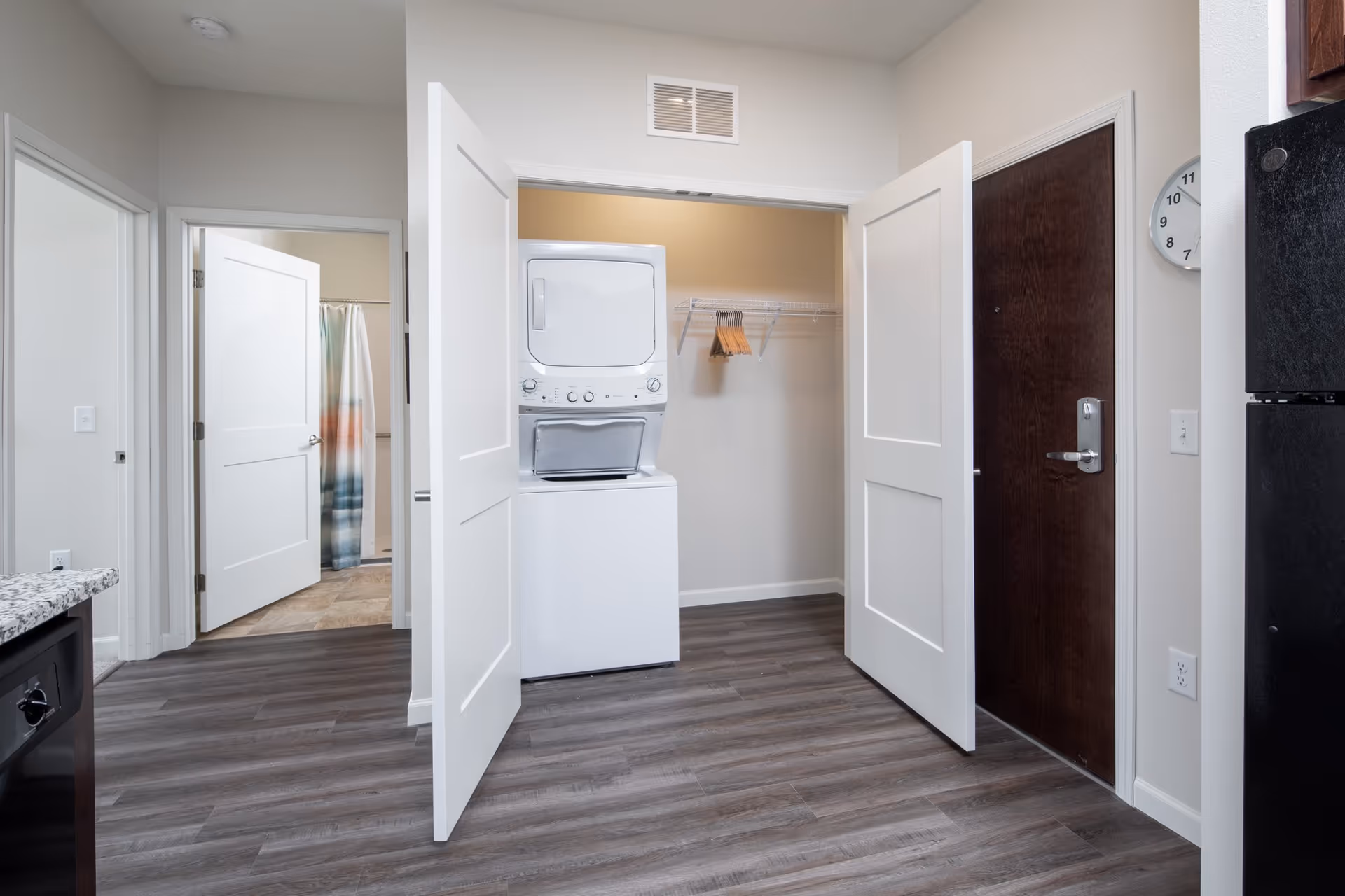 Interior view of a laundry closet with white double doors open, revealing a stacked washer and dryer. To the left, there is a doorway leading to a bathroom with a colorful shower curtain. The floor is covered with dark wood laminate, and a dark wooden entrance door is visible on the right side of the image.