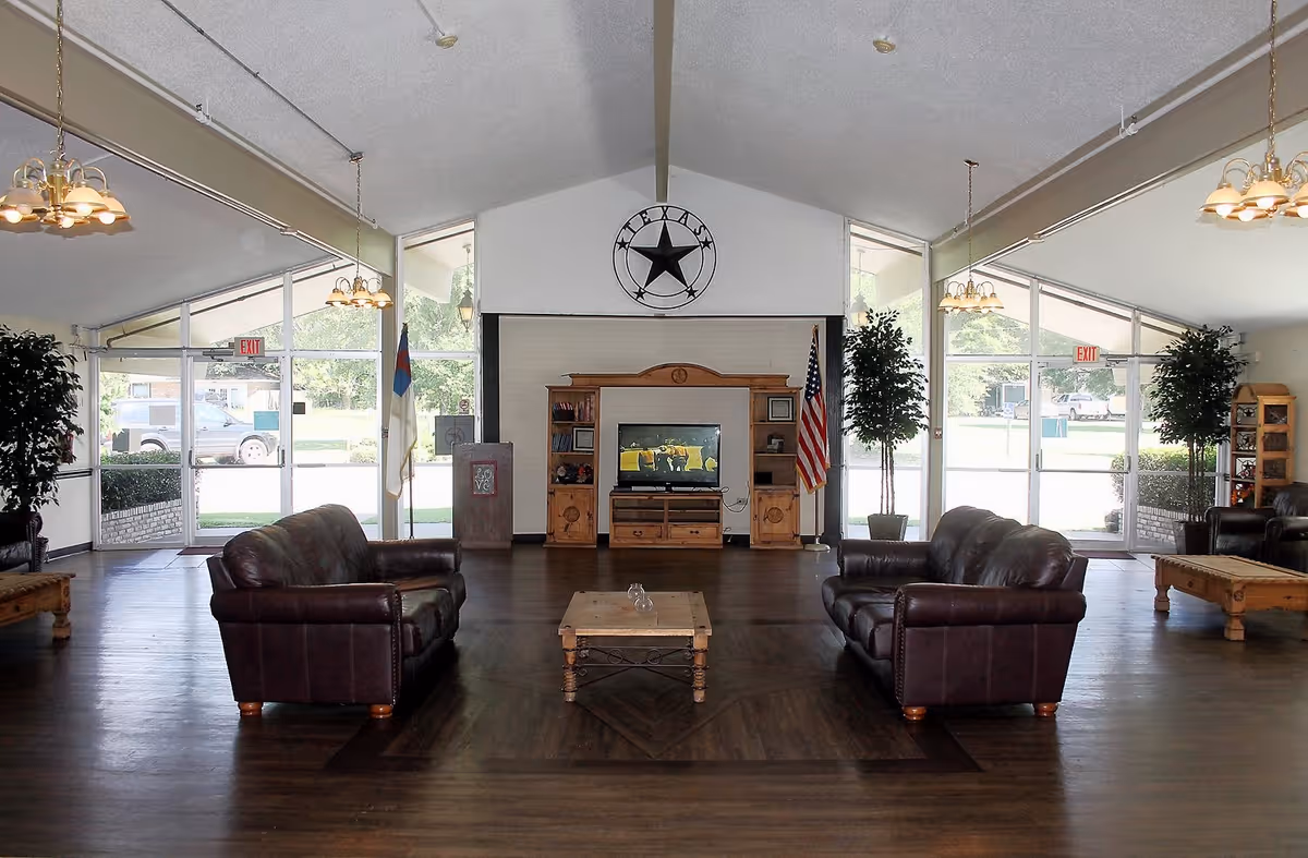 A spacious nursing home common area with two brown leather sofas facing each other across a wooden coffee table. At the far end, there is a wooden entertainment center with a television, flanked by two tall potted plants and two flags. Large windows and glass doors let in natural light, and the ceiling has hanging light fixtures. A large Texas star decoration is mounted on the wall above the entertainment center.