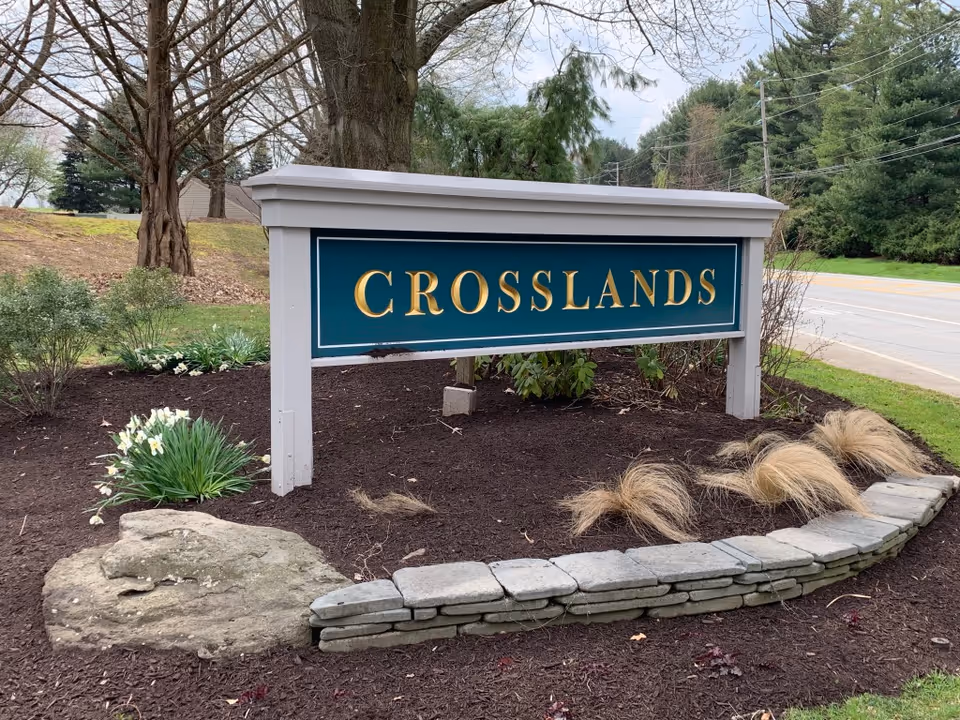 A large outdoor sign with the word 'CROSSLANDS' in gold letters on a green background, mounted on white posts. The sign is surrounded by landscaped soil, plants, and a small stone border, with trees and a road visible in the background.