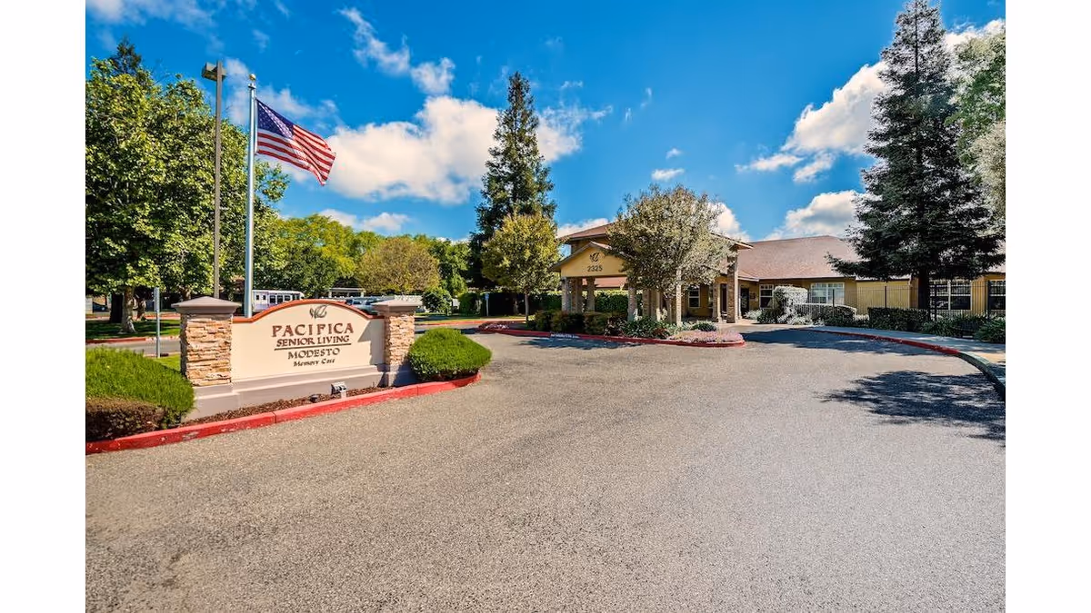 Entrance driveway and sign for Pacifica Senior Living Modesto with an American flag and the facility building under a blue sky.