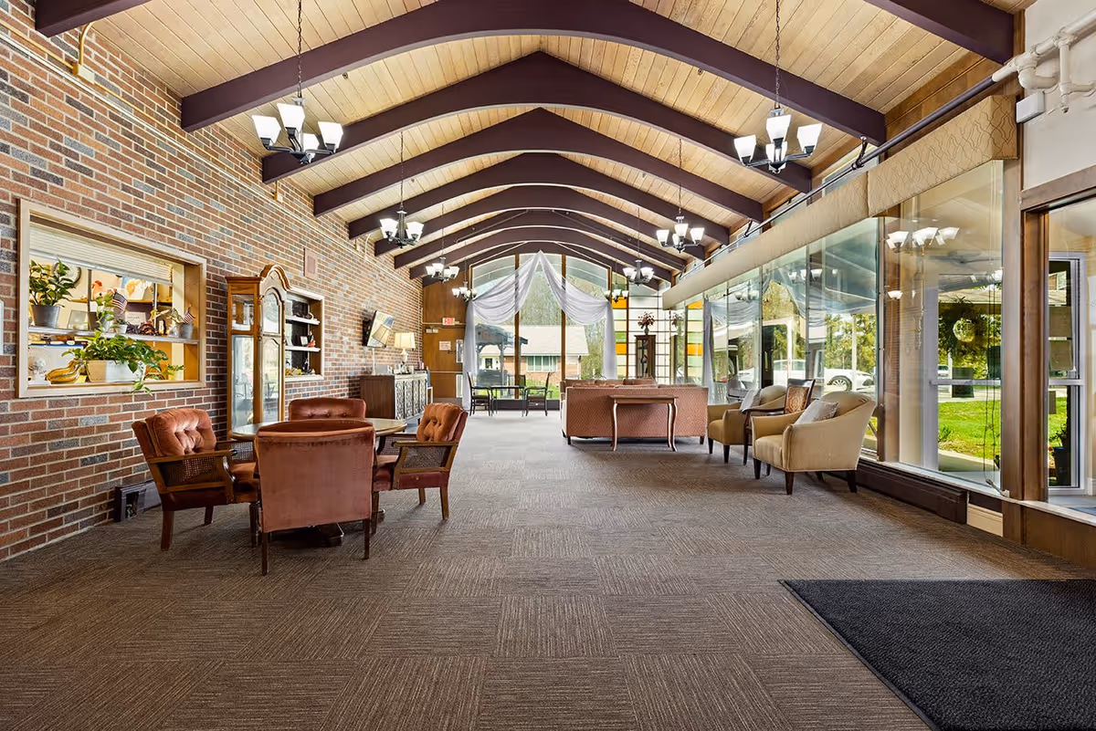 A spacious senior living facility common area with a high vaulted wooden ceiling supported by dark beams. The room features a brick wall on the left with a window opening to another room, decorated with plants and small flags. There are several seating areas with upholstered chairs and sofas arranged around small tables. Large floor-to-ceiling windows on the right side provide a view of the green outdoor area and allow natural light to fill the room. Multiple chandeliers hang from the ceiling, illuminating the space warmly.