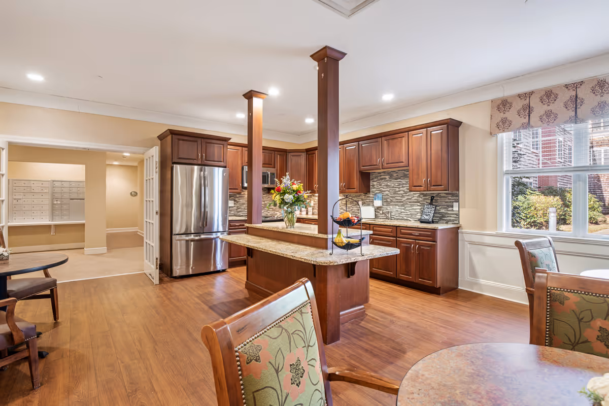 A spacious kitchen area in a senior living facility featuring wooden cabinets, a stainless steel refrigerator, a central island with a granite countertop, and a vase of flowers. The room has wooden flooring and is well-lit with recessed ceiling lights. There are tables and chairs with floral upholstery near large windows that let in natural light. In the background, there is a hallway with mailboxes on the wall.
