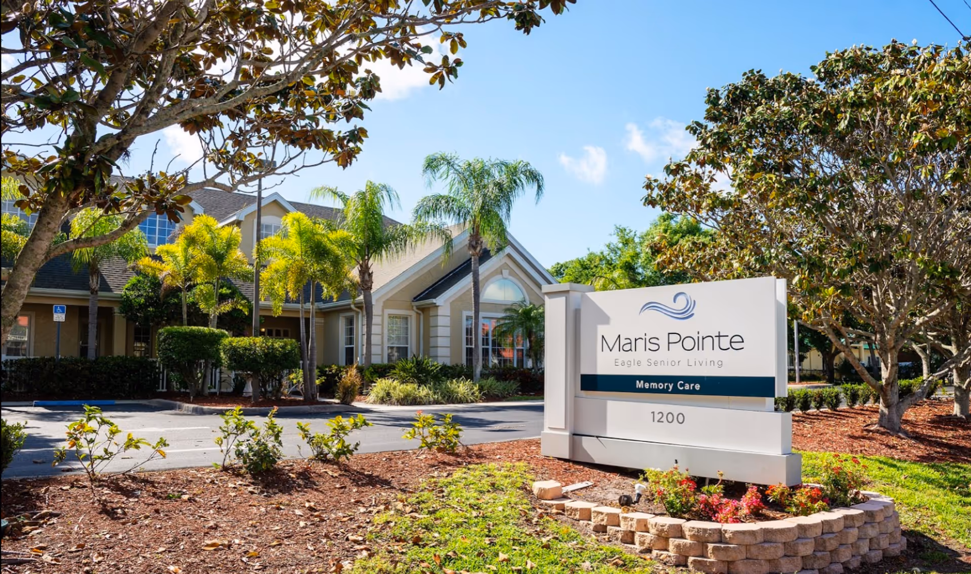 Exterior view of Maris Pointe Eagle Senior Living facility with a sign indicating Memory Care. The building is surrounded by palm trees, bushes, and landscaped grounds under a clear blue sky.