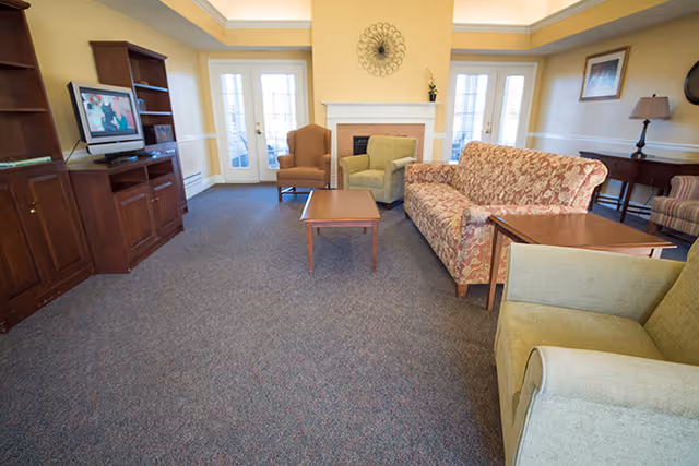 A cozy living room area with a floral patterned sofa, two armchairs, a wooden coffee table, and a fireplace. There is a wooden entertainment center with a TV on the left side and French doors with glass panes in the background letting in natural light. The walls are painted yellow with white trim, and there is a decorative wall piece above the fireplace.