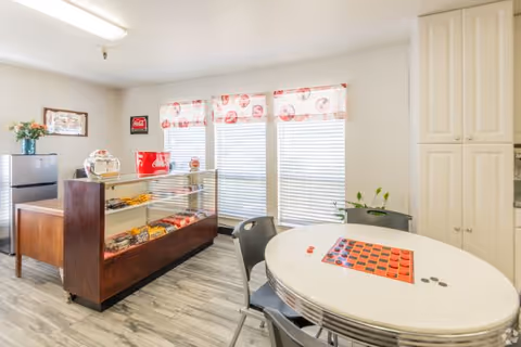 A bright room with large windows covered by white blinds and red patterned valances. There is a round white table with a checkerboard and checkers pieces on it, surrounded by four chairs. To the left, there is a glass display case with snacks and a small refrigerator behind it. The floor has a light wood pattern, and there are white cabinets on the right side of the room.