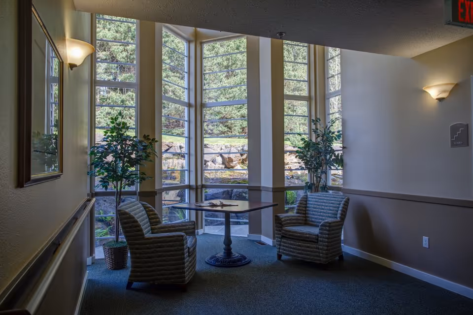 Small sitting area with two upholstered chairs and a table in front of tall windows overlooking landscaped grounds.