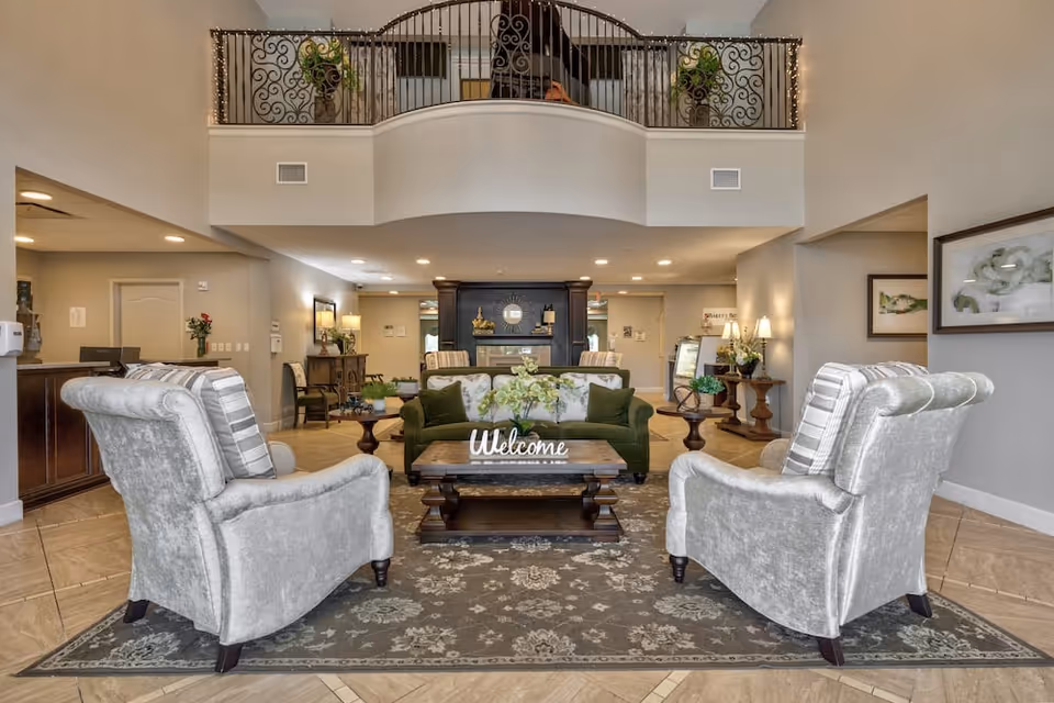 A spacious and elegant living room area in a senior living facility featuring two gray upholstered armchairs with striped pillows, a green sofa, and a wooden coffee table with a 'Welcome' sign and floral arrangement. The room has a high ceiling with a balcony railing above, warm lighting, decorative plants, and framed artwork on the walls.