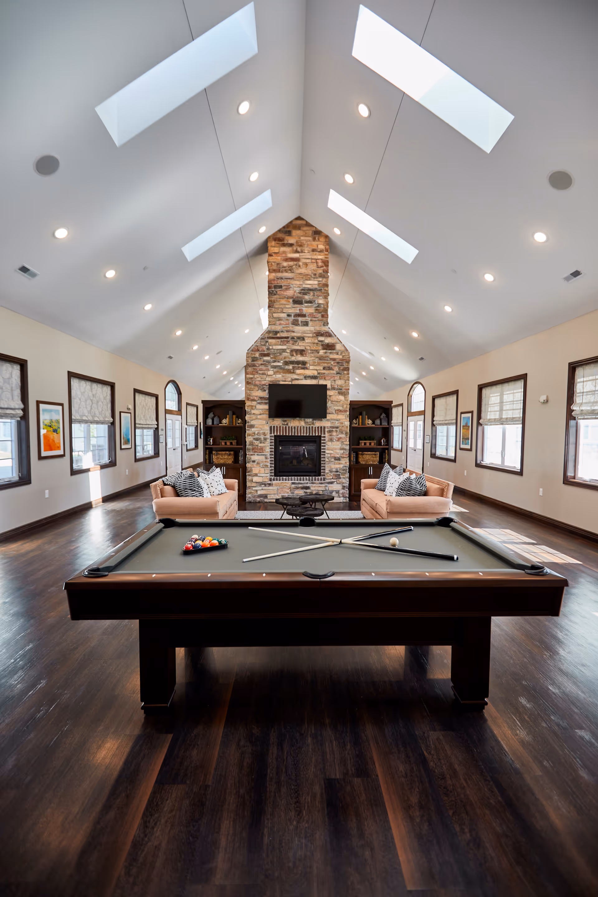 A spacious, well-lit room with a pool table in the foreground, two beige sofas with patterned pillows facing a stone fireplace with a mounted TV above it. The room has high vaulted ceilings with skylights and multiple windows along the walls, wooden flooring, and built-in shelves on either side of the fireplace.