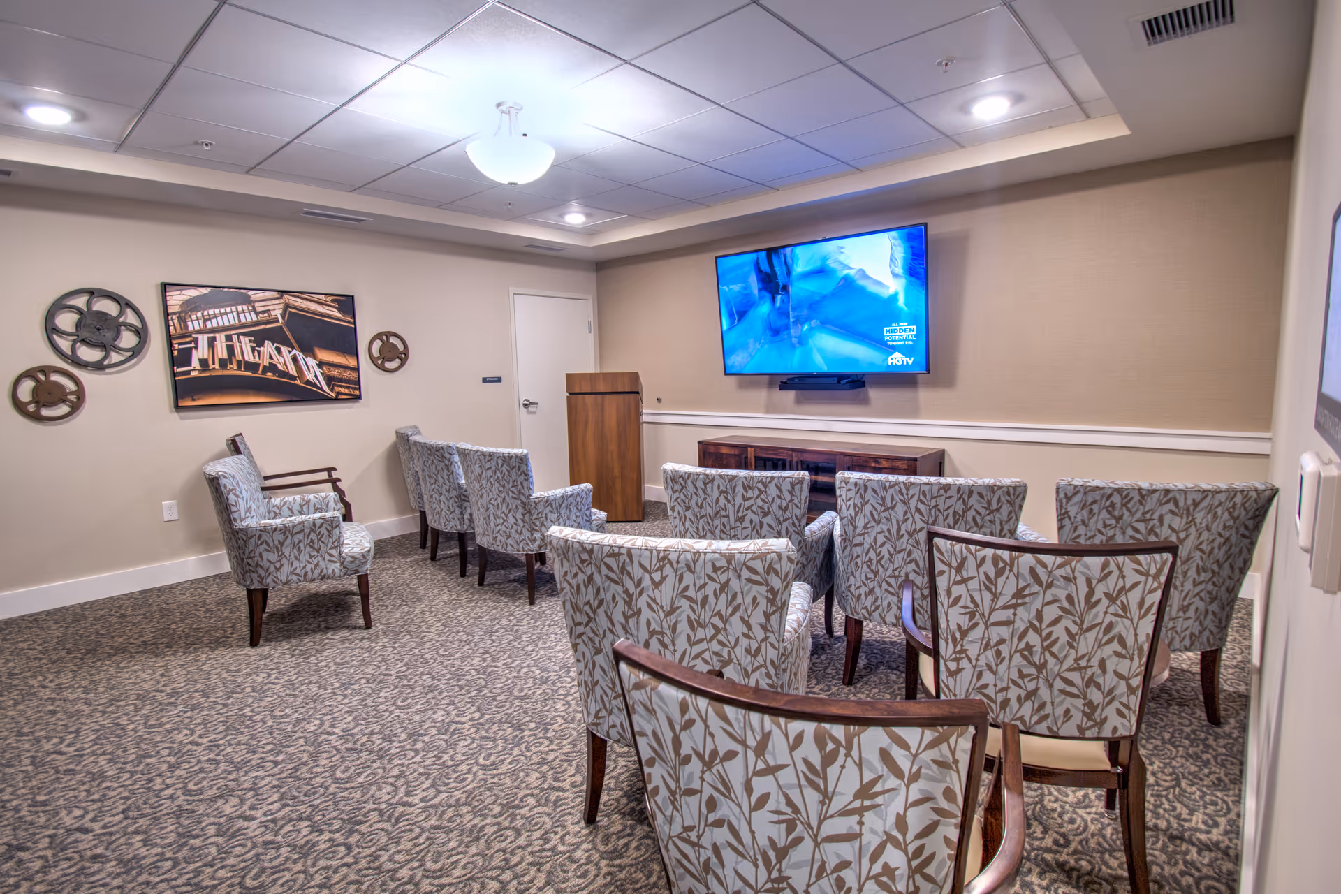 A small theater-style room with patterned armchairs arranged in rows facing a wall-mounted flat screen TV. The room has beige walls, a carpeted floor, and decorative film reels on one wall. There is a wooden podium near the back wall and a door beside it.