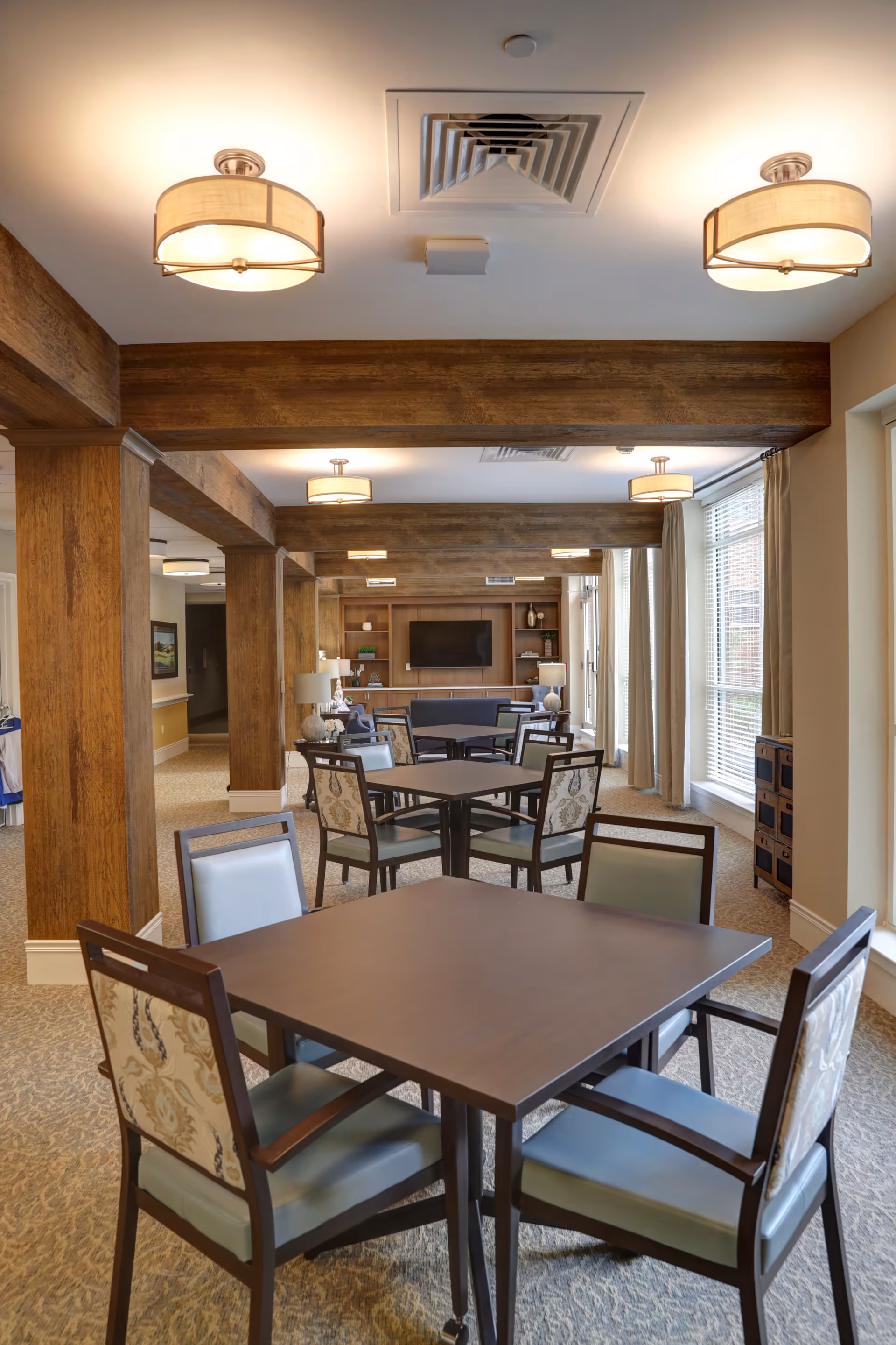 Well-lit communal dining and sitting area with square tables and chairs, exposed wooden beams, and a TV on a built-in shelving wall.