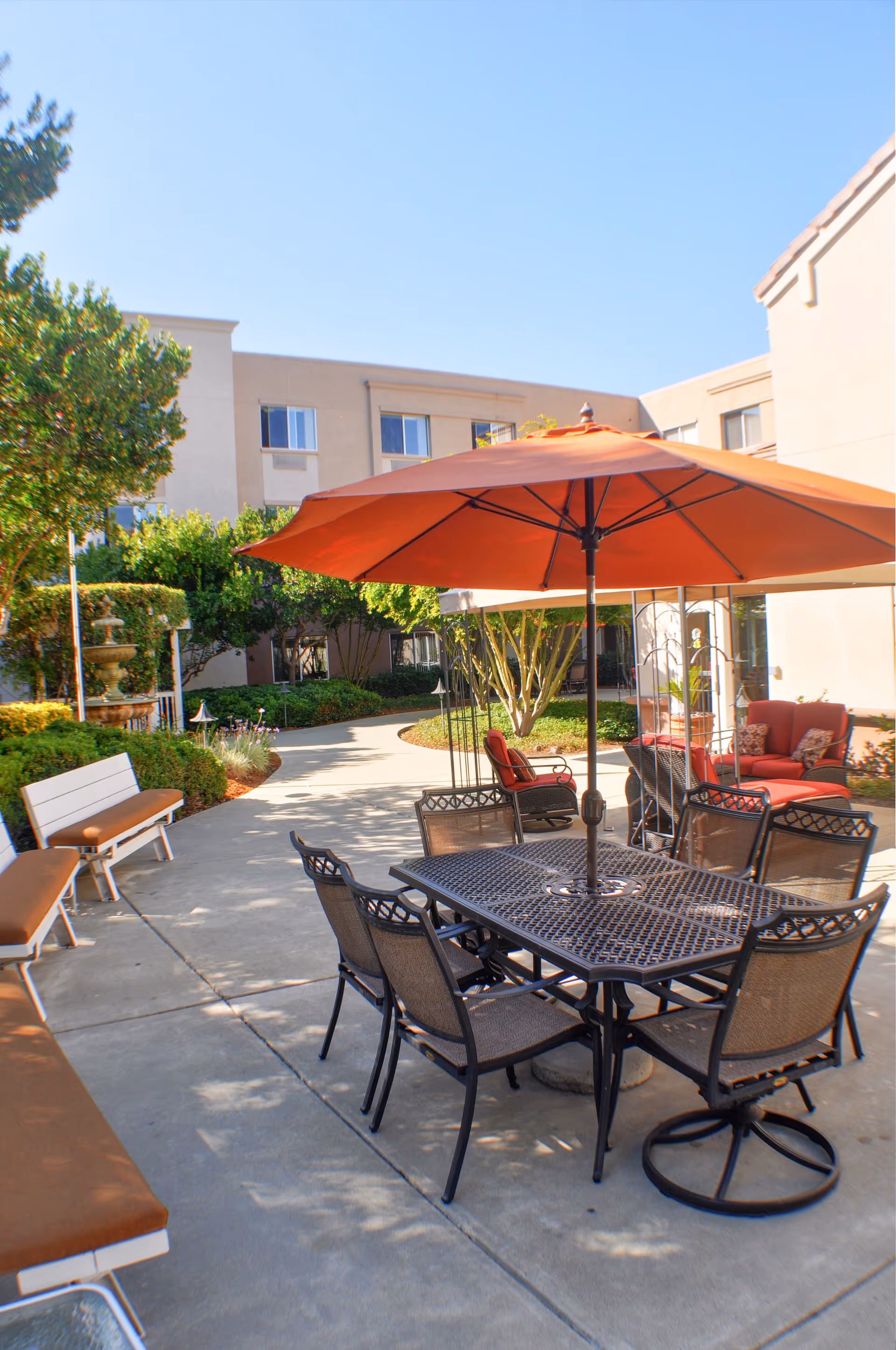 Outdoor senior living courtyard with a metal dining table and chairs under a large orange umbrella, benches and lounge seating near landscaped paths and the building.