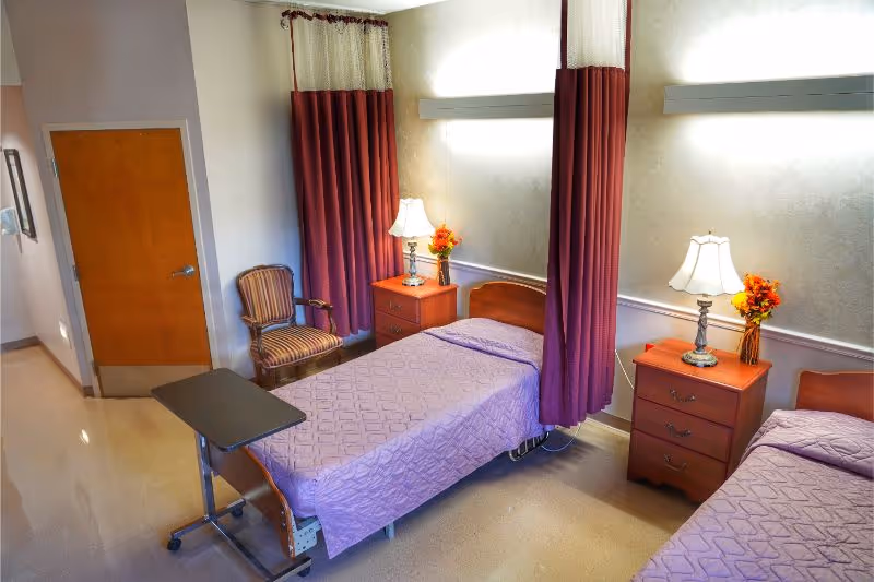A nursing facility room with two single beds covered in purple bedspreads, each accompanied by a wooden nightstand with a lamp and a small vase of flowers. Between the beds are privacy curtains in a matching purple color. There is a wooden chair with striped upholstery and a small overbed table on wheels. The room has a closed wooden door and light-colored walls.