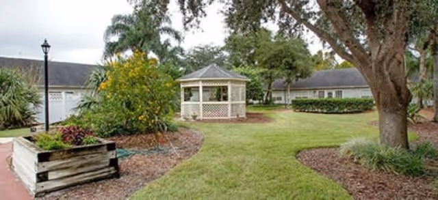 Outdoor garden area at Palmetto Landing featuring a white gazebo in the center, surrounded by green grass, trees, shrubs, and a wooden planter box. Residential buildings are visible in the background under a cloudy sky.