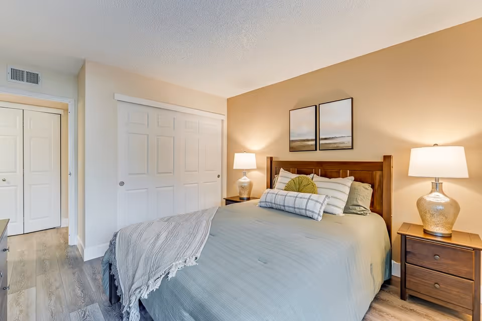 Well-lit bedroom with a made bed, wooden headboard, bedside tables with lamps, framed art above the bed, and sliding closet doors.