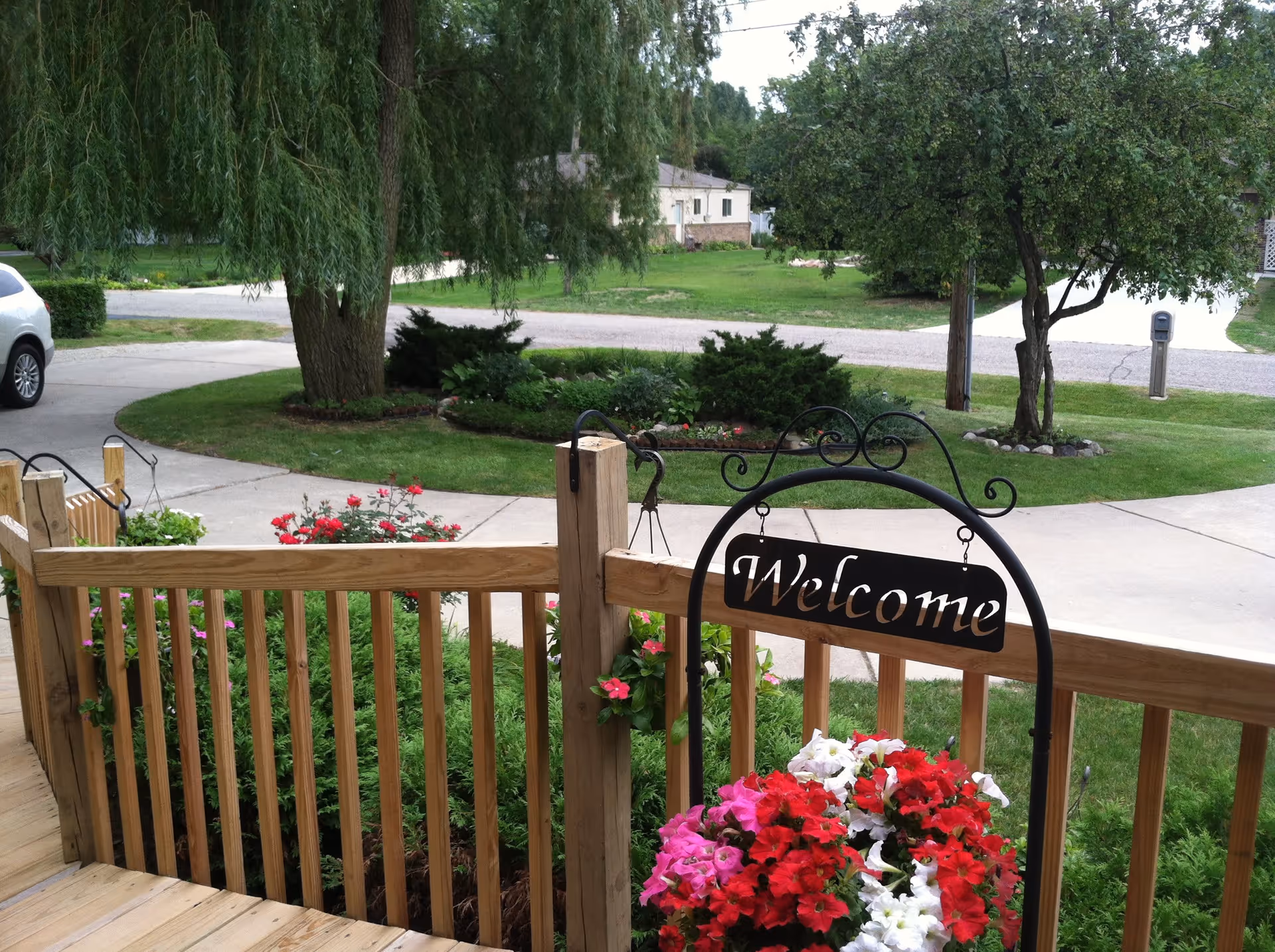 View from a wooden porch with a hanging 'Welcome' sign and a colorful flower arrangement. The porch overlooks a green lawn with trees, shrubs, a driveway, and a parked white car in the background.