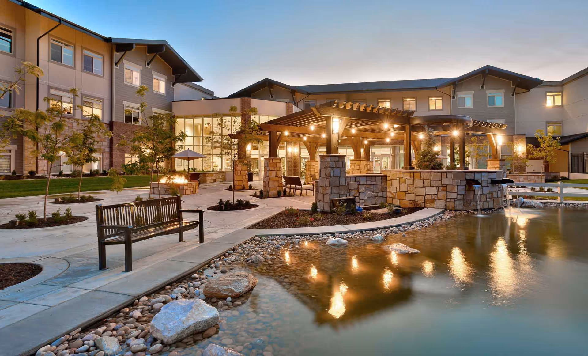 Outdoor courtyard area of Creekside Assisted & Senior Living at dusk, featuring a pond with rocks, a wooden bench, small trees, and a pergola with hanging lights and stone pillars in front of a multi-story building with large windows.
