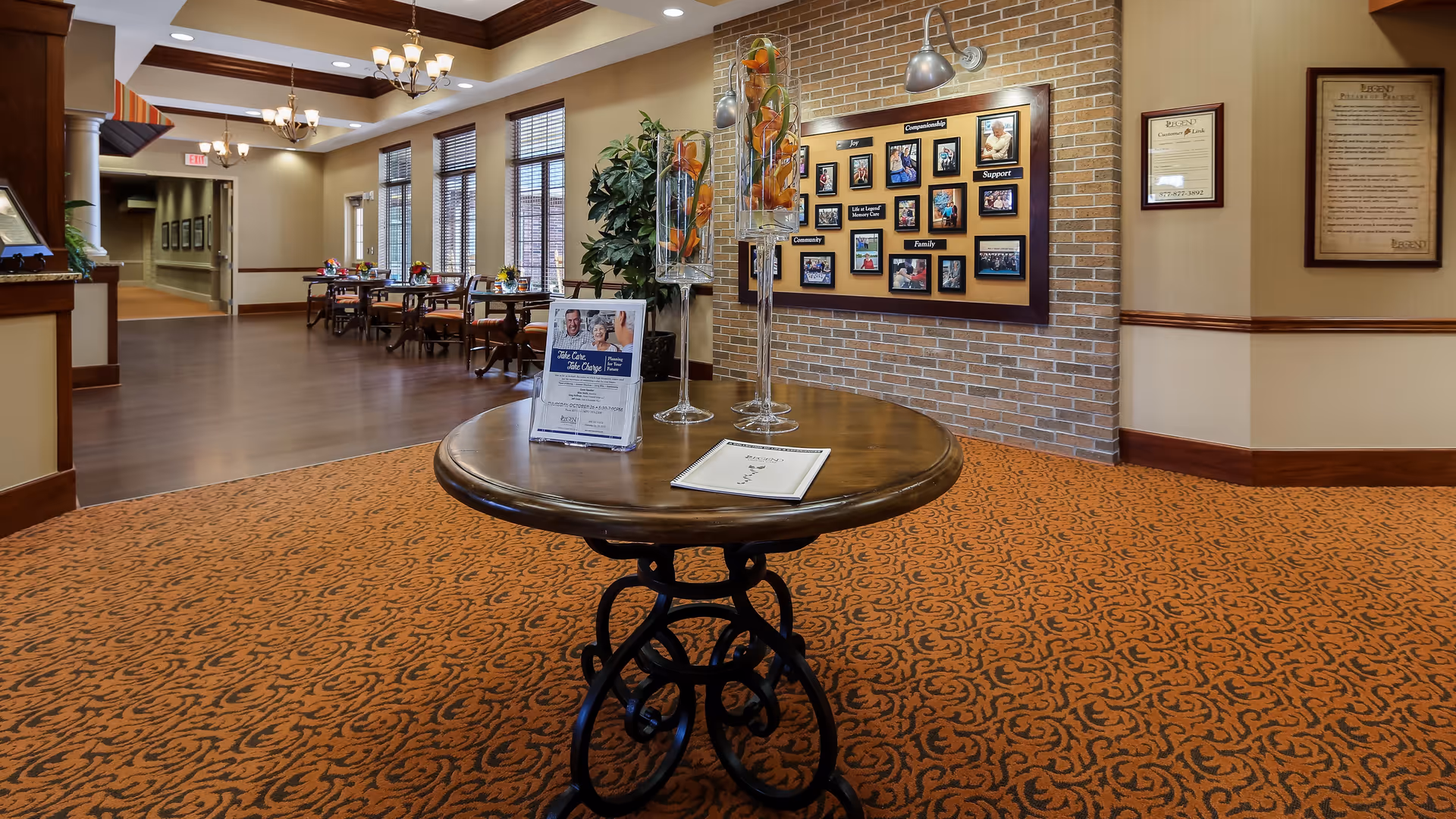 Interior view of a senior living facility with a round wooden table in the foreground displaying decorative glass vases and informational brochures. In the background, there is a dining area with tables and chairs, large windows with blinds, and a brick wall featuring a bulletin board with photos and framed documents on the walls.