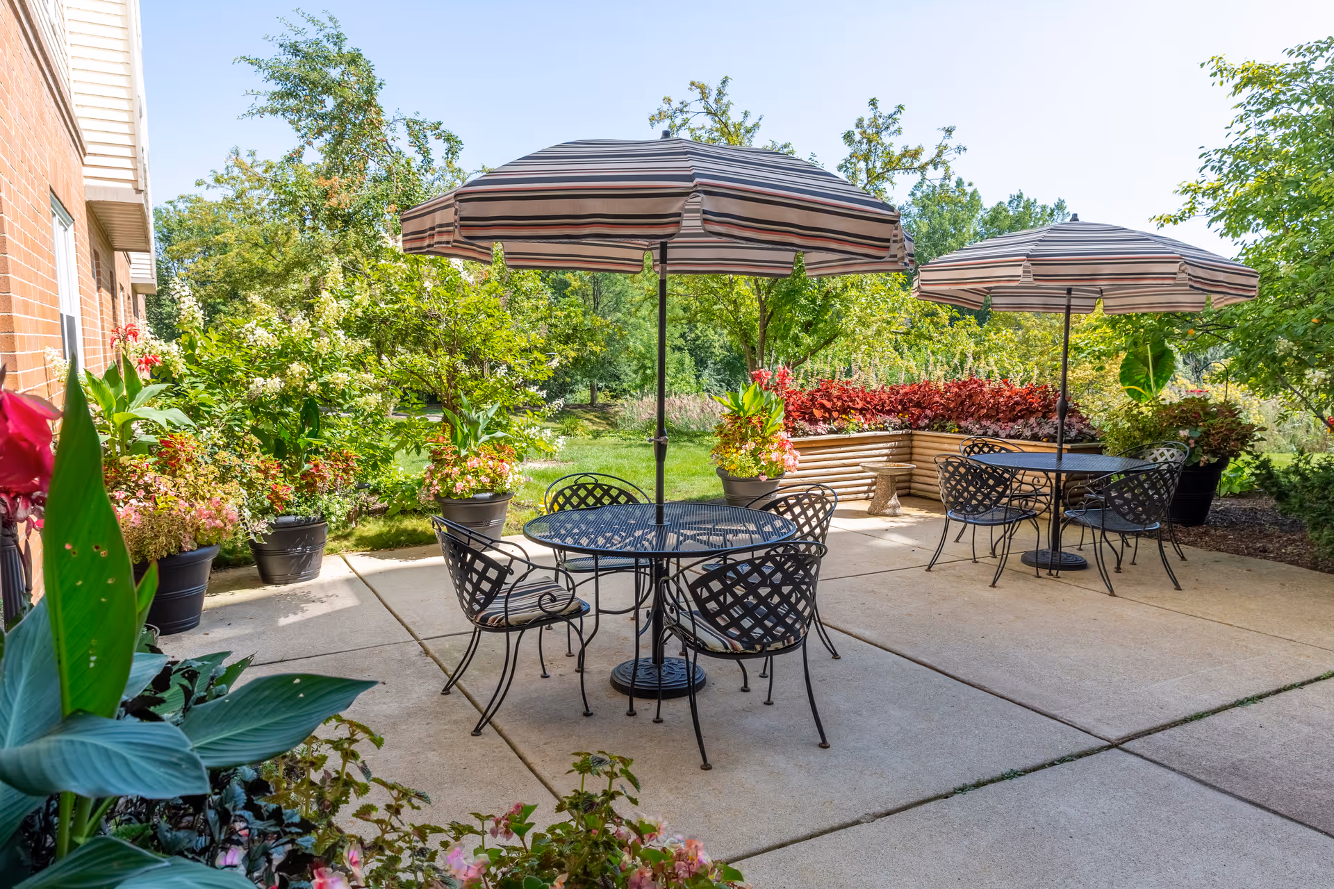 Outdoor patio with metal tables and chairs under striped umbrellas surrounded by potted plants and greenery.