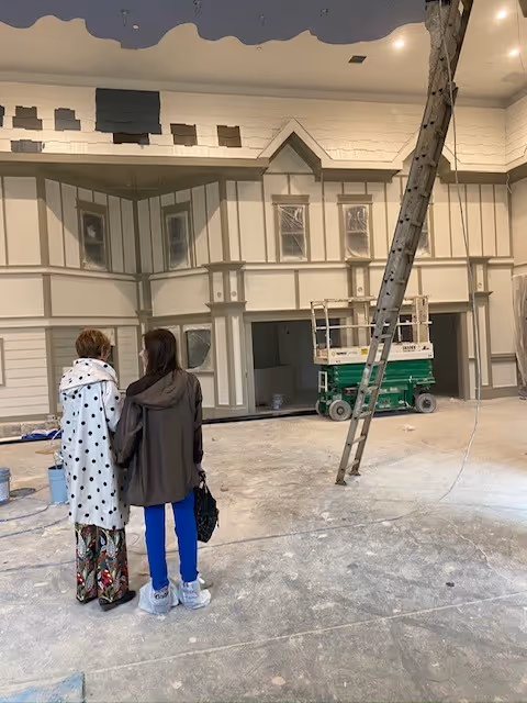 Two people stand inside a large interior under construction, facing a decorative house-like facade with a ladder and a scissor lift.