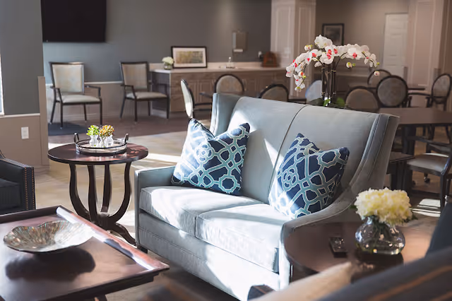 A bright and inviting living room area with a light blue sofa adorned with two patterned blue and white pillows. There are several tables around the sofa, including a round side table with small plants and a coffee table with a decorative bowl. In the background, there are additional chairs and a counter with framed artwork and a flower vase. The room is well-lit with natural light.