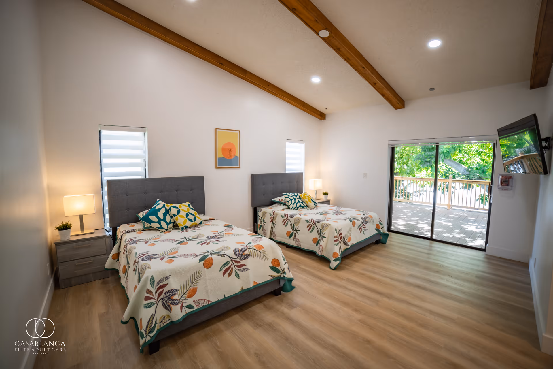 A bright bedroom with two beds featuring floral-patterned bedspreads and colorful pillows. Each bed has a gray upholstered headboard and a nightstand with a lamp and small plant. The room has wooden beams on the ceiling, light wood flooring, two small windows with blinds, a framed artwork on the wall, and a sliding glass door leading to an outdoor balcony with greenery visible outside. A wall-mounted TV is on the right side of the room.