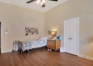 A simple bedroom with a single bed, walker, nightstand with lamp, double closet doors, ceiling fan, and hardwood floor.