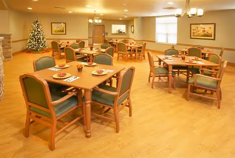 A dining room with several wooden tables and green cushioned chairs arranged neatly. Each table is set with plates, napkins, and utensils. There is a decorated Christmas tree in the corner, light wood flooring, and framed artwork on the walls. The room is well-lit with ceiling lights.