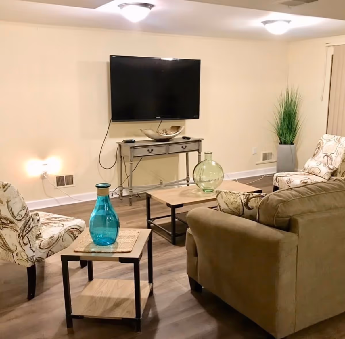 A cozy living room with a mounted flat-screen TV on a beige wall above a small wooden console table. The room features a brown sofa, two patterned armchairs, a wooden coffee table with a green glass vase, and a side table with a blue glass vase. There is a tall green plant in a gray pot in the corner, and the floor is covered with wood-like flooring.