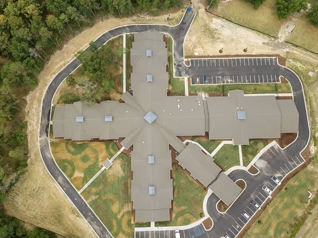 Aerial view of Lakeside Place Senior Living facility showing a large building with a cross-shaped layout surrounded by parking lots, driveways, and green landscaped areas with trees around the perimeter.