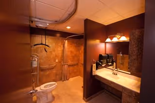 Interior view of a bathroom in a senior living facility featuring a toilet with grab bars, a walk-in shower with a curtain, a sink with a countertop, a mirror, and wall-mounted soap dispensers. The walls and floor have a warm brown tone, and the lighting is soft and warm.