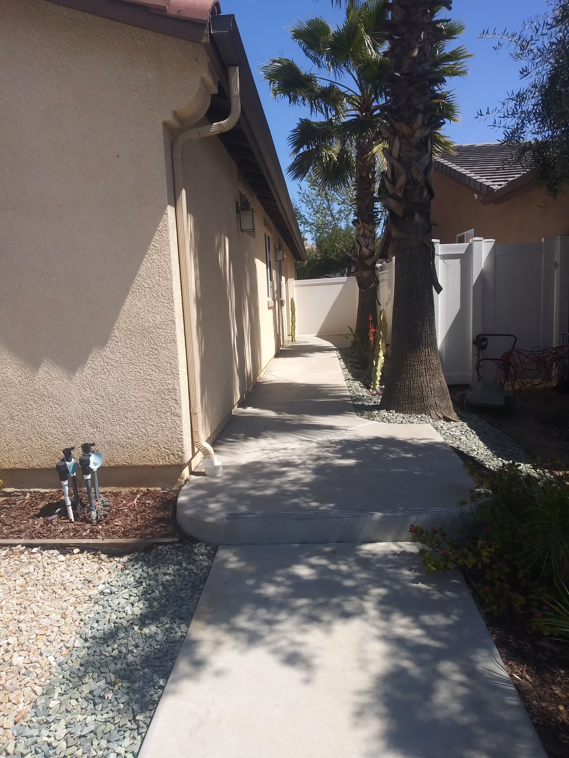 Concrete side walkway along a stucco house with palm trees and a white fence in the background.