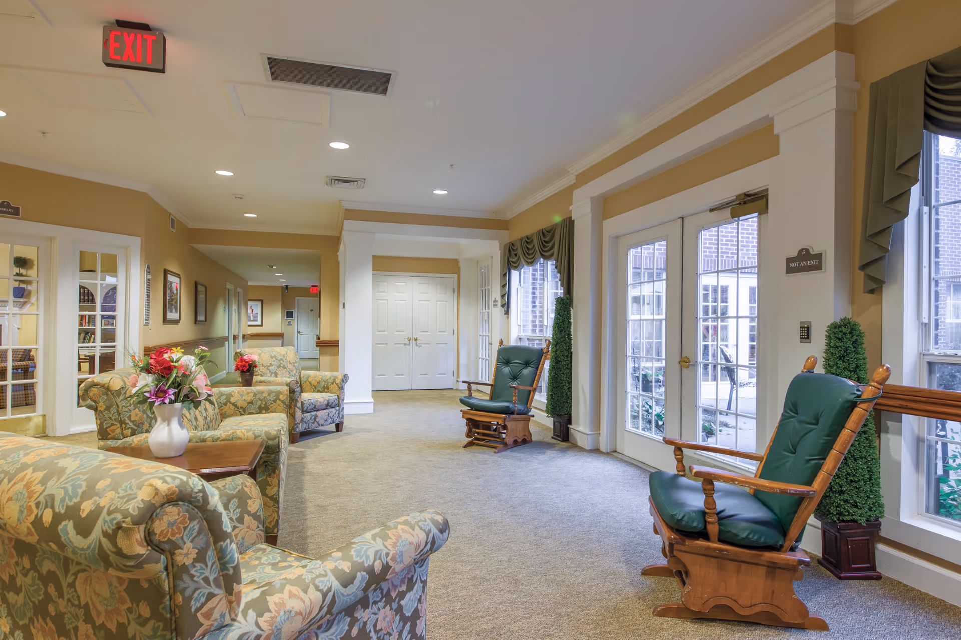 A bright and spacious common area in a senior living facility with floral upholstered armchairs and wooden rocking chairs with green cushions. There are large windows with green drapes letting in natural light, a vase with flowers on a wooden side table, and a hallway leading to other rooms. An exit sign is visible on the ceiling.