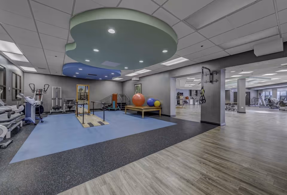 A spacious fitness and rehabilitation room in a senior living facility featuring exercise equipment, parallel bars for walking practice, large colorful exercise balls on a wooden bench, and a ceiling with decorative circular panels and recessed lighting. The room has a mix of blue, black, and wood-patterned flooring and opens into a larger gym area with additional exercise machines.