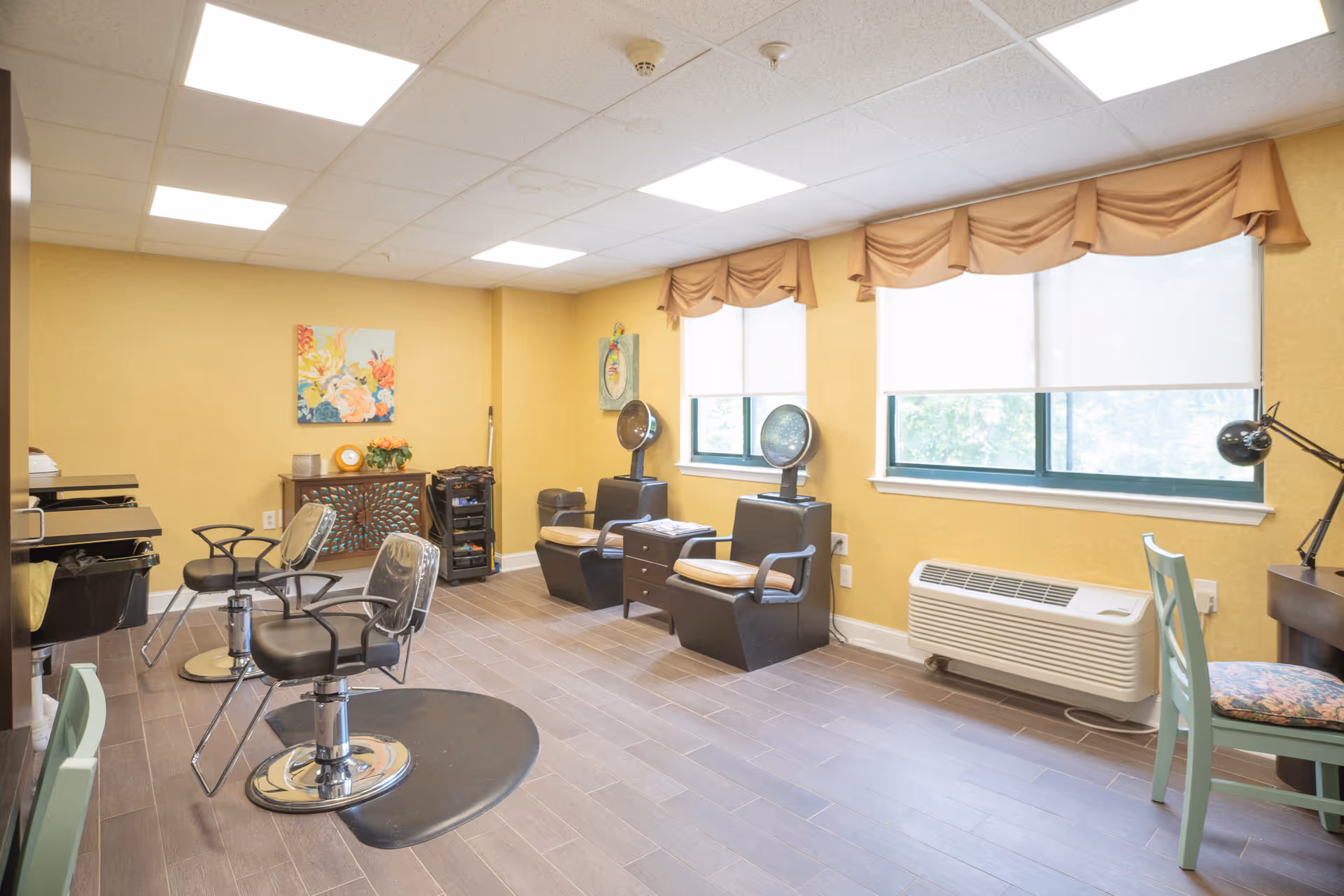 Bright salon room with styling chairs, hooded hair dryers, sinks and large windows in a senior living facility.