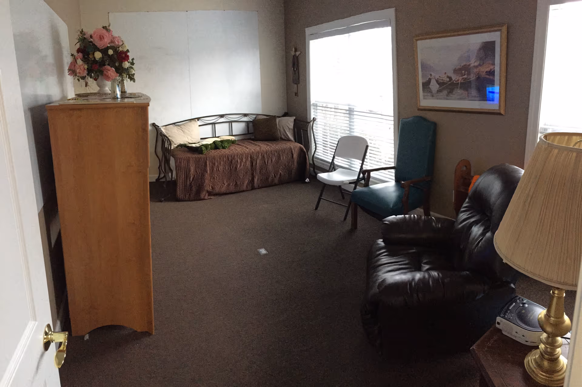 A cozy living room with a brown carpet, a metal-framed daybed with pillows and a brown cover, a wooden dresser with a vase of flowers on top, a white folding chair, a teal armchair, a dark brown leather recliner, a side table with a brass lamp and a CD player, and a framed picture on the wall. Two large windows with blinds allow natural light into the room.