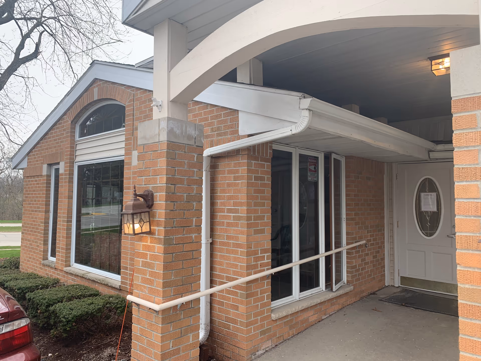 Covered brick building entrance with large windows, a white oval-paneled door, and a wall-mounted lantern.
