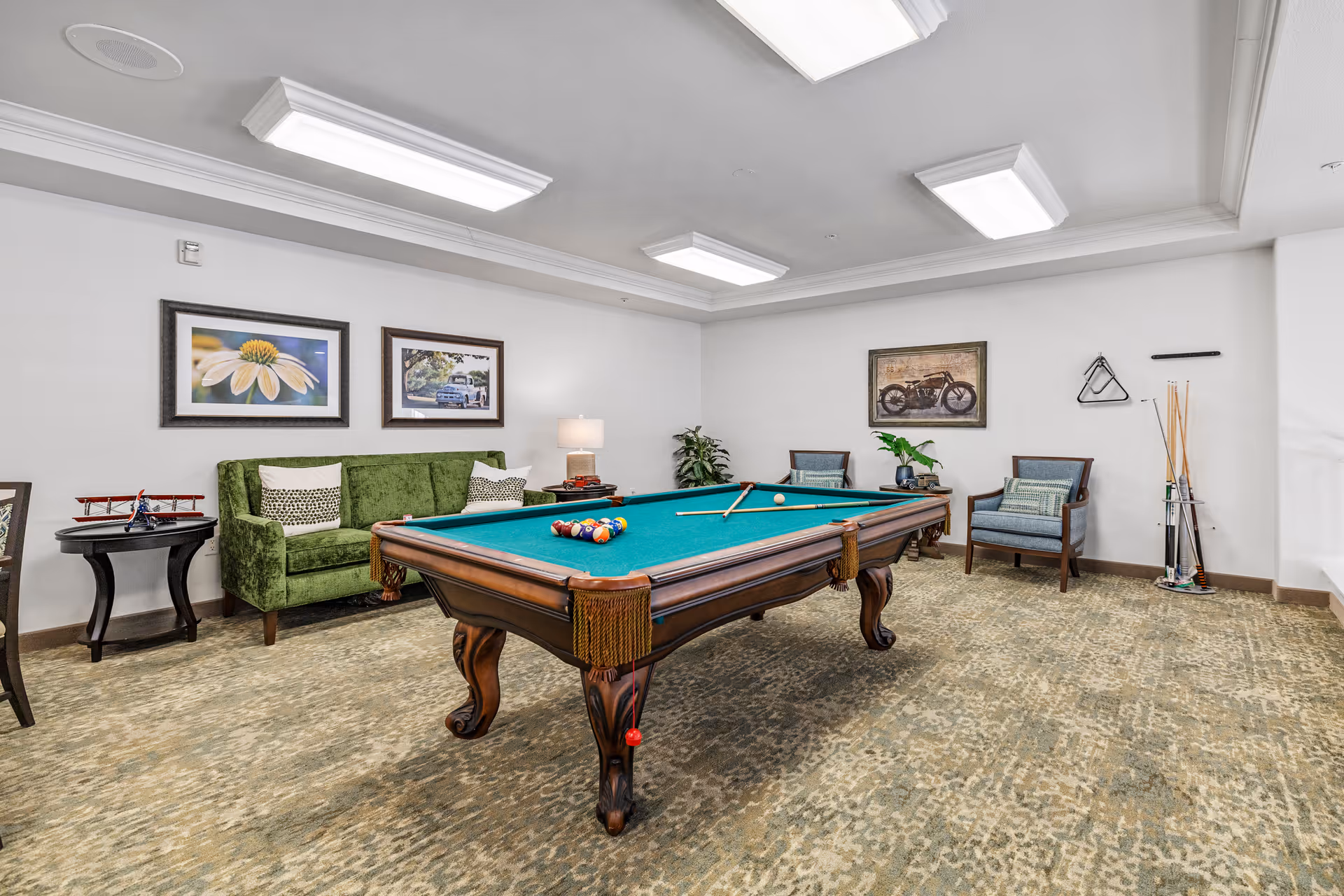 A recreational room with a green felt pool table in the center, surrounded by a green sofa, two blue armchairs, a side table with a model airplane, and framed artwork on the walls. The room has carpeted flooring and bright ceiling lights.