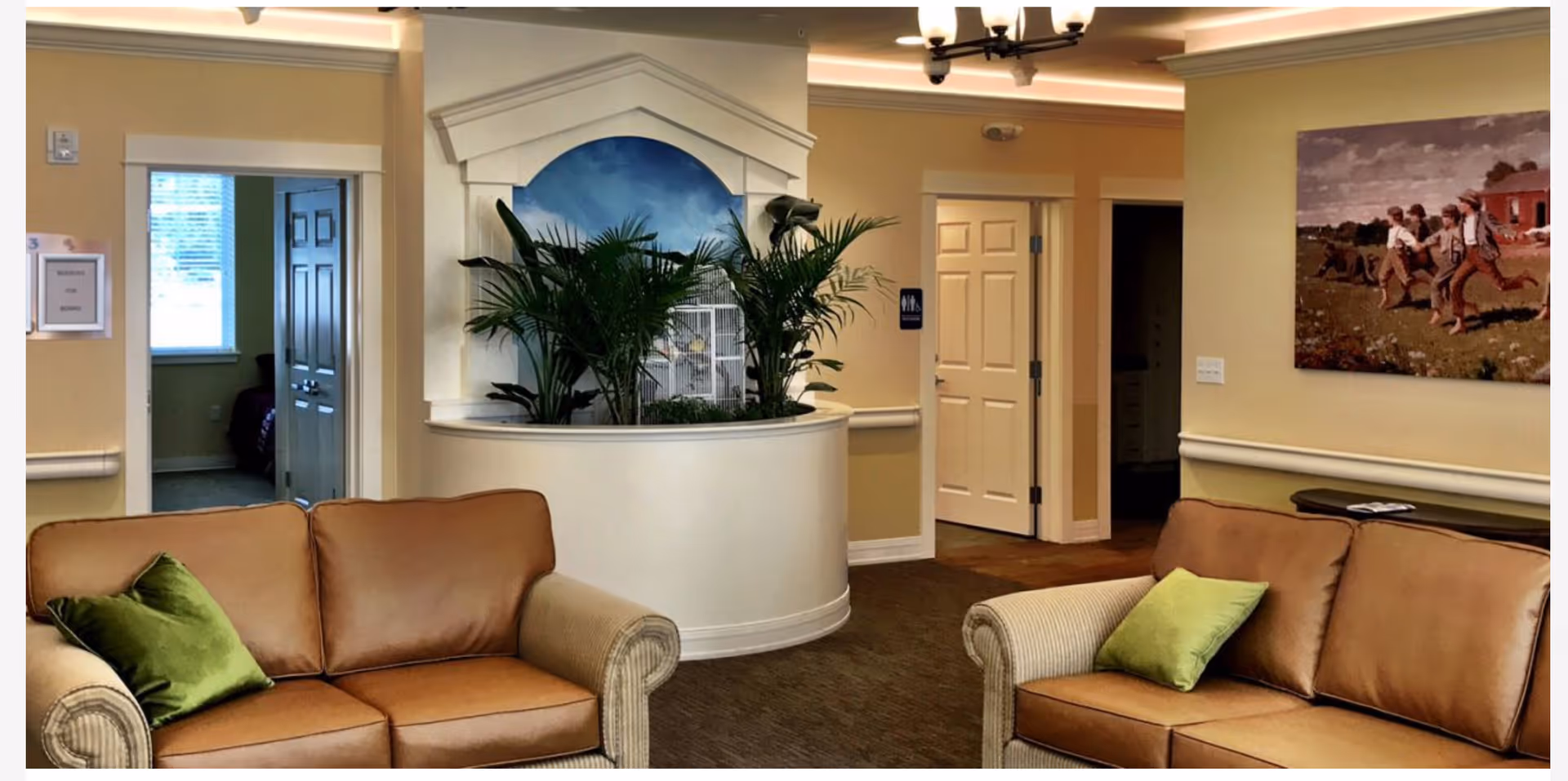 A cozy interior common area with two brown leather sofas each with a green pillow, a curved white planter with green plants, beige walls, and doorways leading to other rooms. There is a painting on the wall depicting children playing outdoors.