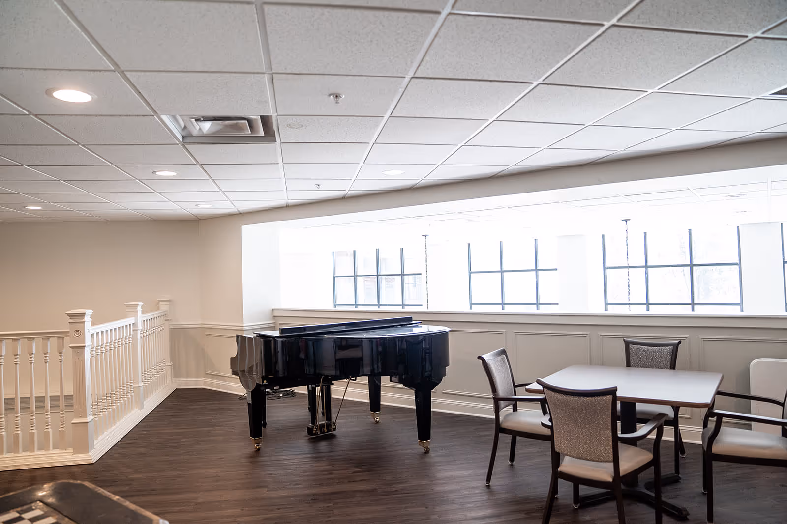 Interior room with a black grand piano positioned near a large window with multiple panes. To the right of the piano, there is a square table surrounded by four cushioned chairs. The room has dark wood flooring, white paneled walls, and a white ceiling with recessed lighting. A white railing is visible on the left side of the image.