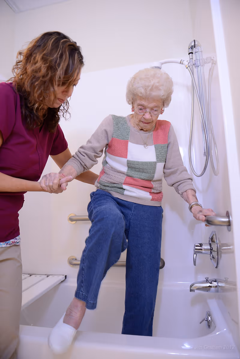 A caregiver helps an elderly woman step into a bathtub in an accessible bathroom.