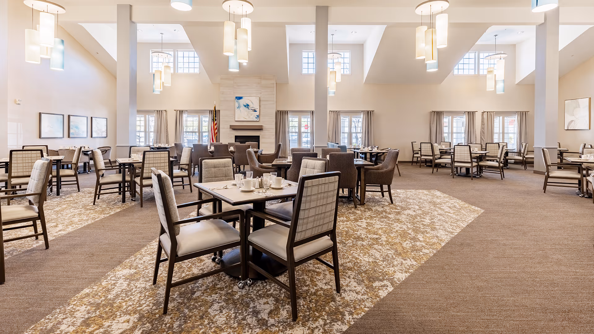 A spacious dining room with multiple tables and chairs arranged neatly. The room features high ceilings with modern hanging light fixtures, large windows with curtains, and a fireplace with artwork above it. The carpet has a patterned design in the center area where the tables are placed.
