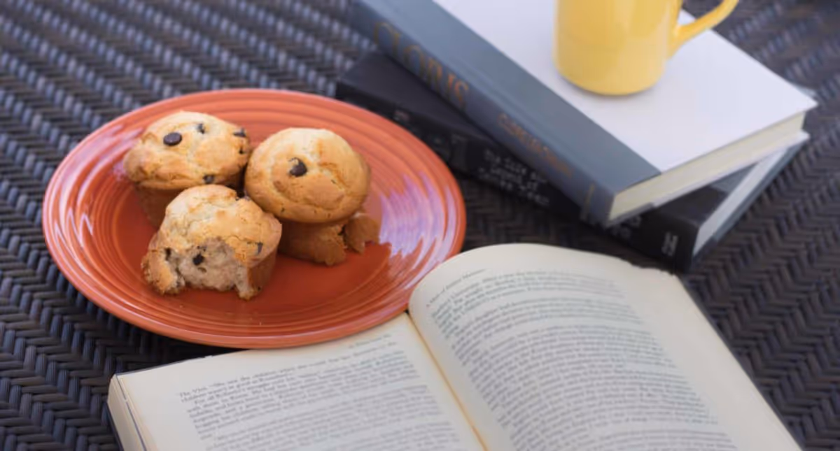 Close-up of a red plate with three chocolate chip muffins, one partially eaten, placed on a woven surface next to an open book and a stack of closed books with a yellow mug on top.